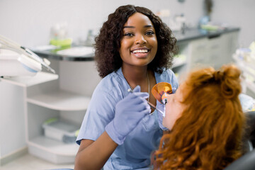 Smiling black woman dentist wearing blue uniform, providing tooth restoration and filling with curing polymerization UV lamp for her little patient, cute girl with red curly hair - Powered by Adobe