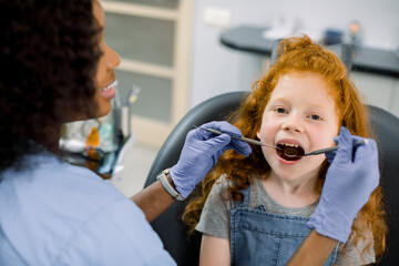 A child at dental clinic. Close up portrait of little child girl with red curly hair, sitting with open mouth in dental chair while female African dentist makes teeth check up using dental mirror.