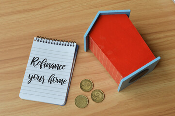 Top view of toy wooden house, coins and notebook written with text REFINANCE YOUR HOME.