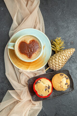 Above view of a cup of frothy coffee on towel with accessory and small cupcakes with chocolates on dark background