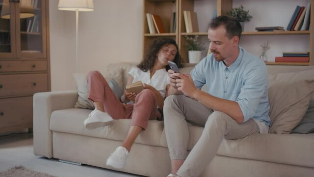Medium shot of unhappy couple sitting on sofa man having affair with somebody else texting messages on smartphone and woman feeling jealous starting fighting and quarrelling