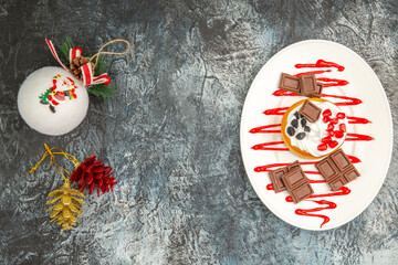 top view berry cake and chocolates on white oval plate white xmas tree ball and colorful pinecones on grey-white background with copy space