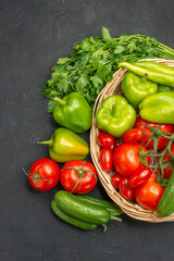 Vertical view of fresh vegetables red tomatoes with stems green peppers and cucumbers inside and outside of basket on dark background