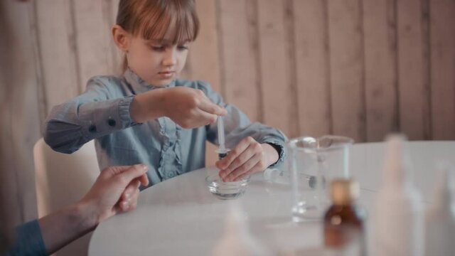 Slow-motion medium close-up of little girl flipping hydrogen peroxide into syringe and adding it into measuring cup with ethanol for making antiseptic from covid-19
