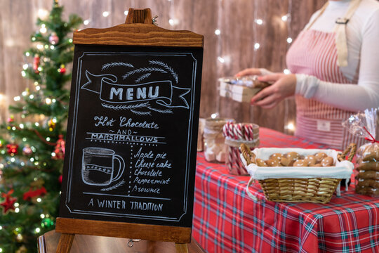 Christmas pastry shop . Woman seller, waitress selling gingerbread, marshmallows, cookies, sweets in a small cozy cafe. Homemade bakery menu in the foreground