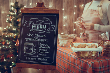 Christmas pastry shop . Woman seller, waitress selling gingerbread, marshmallows, cookies, sweets in a small cozy cafe. Homemade bakery menu in the foreground