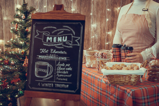 Christmas pastry shop . Woman seller, waitress selling gingerbread, marshmallows, cookies, sweets and hot chocolate in a small cozy cafe. Homemade bakery menu in the foreground