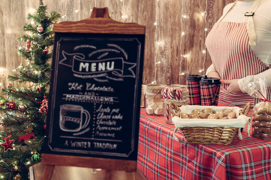 Christmas pastry shop . Woman seller, waitress selling gingerbread, marshmallows, cookies, sweets and hot chocolate in a small cozy cafe. Homemade bakery menu in the foreground