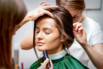 The hairdresser and makeup artist are doing hairstyle and make-up for a beautiful young caucasian...