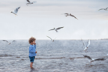 A kid with curly hair in a sailor's striped long sleeve shirt feeds gulls on the sea. Cloudy weather, vintage toning.