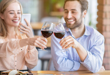 Young woman making photo of romantic toast with clinking wine glasses during festive dinner at cafe, selective focus