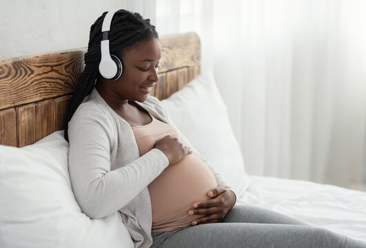 Happy black pregnant woman lying in bed, listening music in wireless headphones