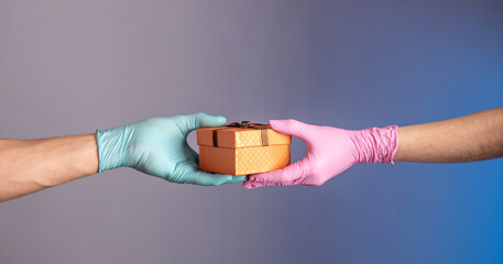 A woman's hand in pink glove is giving a gift box to man's hand in a blue glove a on a blue and gray background. Concept of a lifestyle in a pandemic