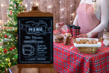 Christmas pastry shop . Woman seller, waitress selling gingerbread, marshmallows, cookies, sweets and hot chocolate in a small cozy cafe. Homemade bakery menu in the foreground