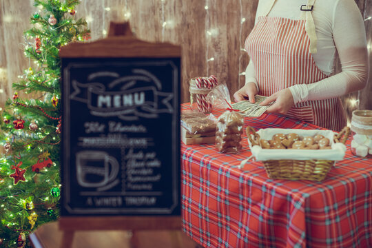 Christmas pastry shop. Woman seller, waitress counting money selling gingerbread, marshmallows, cookies, sweets in a small cozy cafe. Homemade bakery menu in the foreground