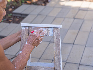 Senior man holding a brush to paint white on the old wooden chair without a backrest. Old chairs...