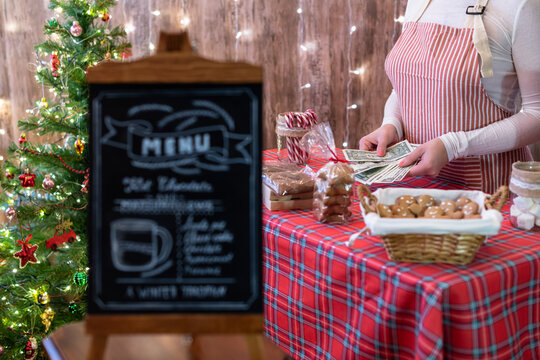 Christmas pastry shop. Woman seller, waitress counting money selling gingerbread, marshmallows, cookies, sweets in a small cozy cafe. Homemade bakery menu in the foreground