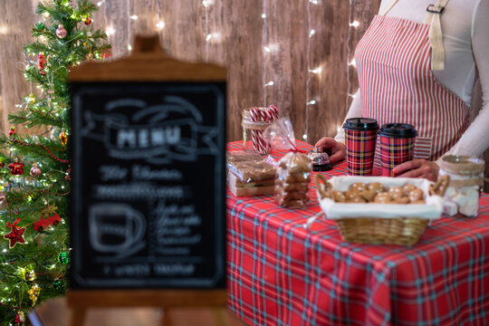 Christmas pastry shop . Woman seller, waitress selling gingerbread, marshmallows, cookies, sweets and hot chocolate in a small cozy cafe. Homemade bakery menu in the foreground - Powered by Adobe