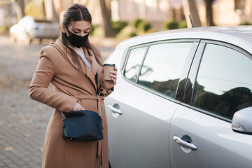 Woman in black face mask hold cuf of coffee by the car and look car key. Woman open car. Protectiv mask in quarantine. Cov-19