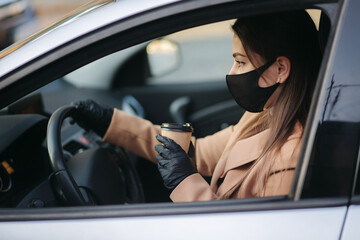 Fototapeta na wymiar Woman with face mask driving her car with cup of coffee during coronavirus pandemic, Covid-19