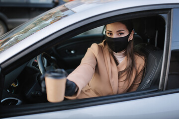 Naklejka premium Attractive young woman take cup of coffe while sitting in car. Woman in face mask and gloves. Coffee blured