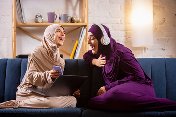 Laughting. Happy and young two muslim women at home talking, smiling, having fun, sitting on sofa....