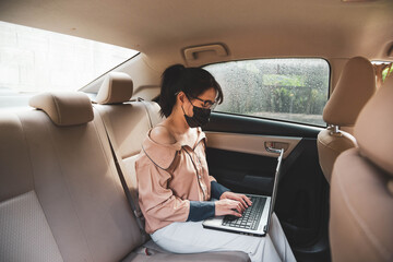 working women sitting in the back of a car wearing a protective mask and working at a computer