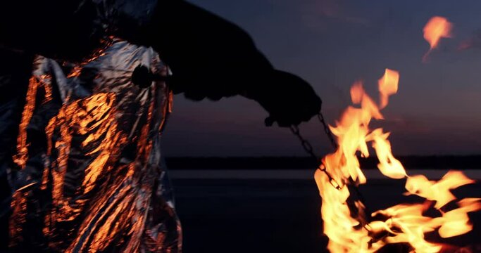 A fire spinner in a silver suit carries torches burning in flames on chains, late at night. Preparing for the fire show.