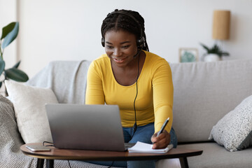 Smiling black lady in headset writing notes while watching webinar on laptop