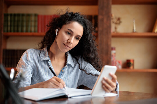 Woman sitting at desk, using smartphone and writing in notebook