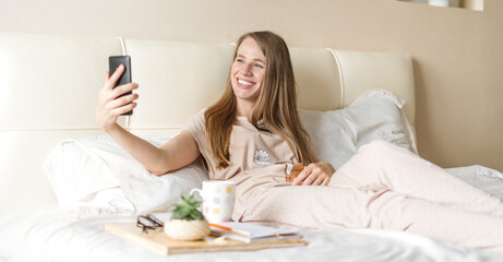 Obraz na płótnie Canvas Happy, smiling, relaxed, and cheerful young woman taking a selfie on smartphone while sitting on bed with coffee. Blonde girl uses cellphone and having video call. Enjoying free leisure time at home.