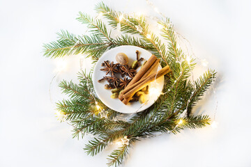 spices for mulled wine on a small white plate on a white background with green branches of a Christmas tree and a burning garland of warm yellow color. the view from the top