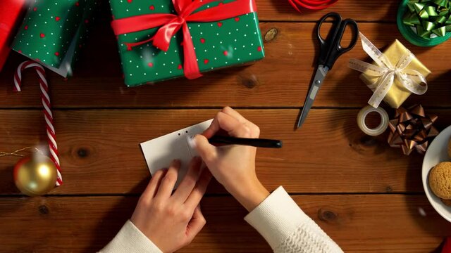 holidays, new year and christmas concept - hands packing gift box, attaching tag and writing receiver name at home over snow