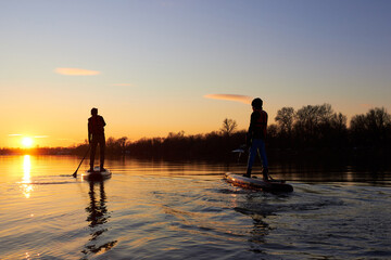 Silhouettes of two boys on stand up paddle board at dusk on a flat quiet winter river with beautiful sunset colors