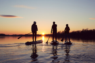Silhouettes of the people on stand up paddle board at dusk on a flat quiet winter river with...