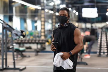 Black bodybuilder in face mask holding water, towel at gym