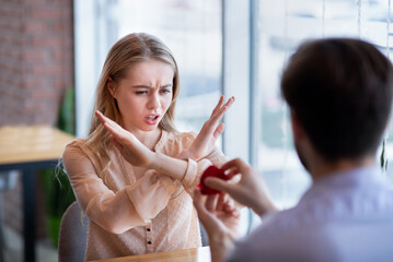 Scared young woman rejecting marriage proposal, gesturing NO, refusing to accept engagement ring at...