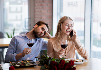 Young woman speaking on mobile phone during dull date, her boyfriend feeling lonely and bored at...