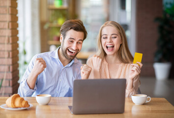 Cheerful Caucasian couple with credit card and laptop shopping online at city cafe