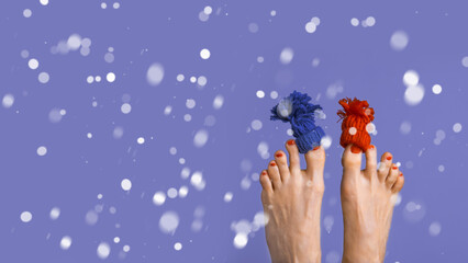 Funny playful female feet with small knitted hat on big toes over purple color background with falling snow