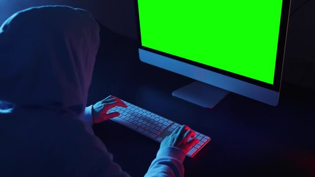 Back view of hacker programmer wearing a hood, typing very fast intelligible code in a computer. Unrecognizable woman hacking in a dark room. Green screen display for chroma key mock up. 
