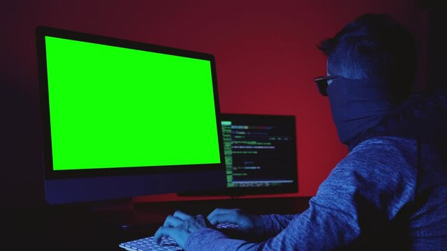 Man with a covered face in front of two computer screens. Criminal hacker programming intelligible codes  Computer crime concept. Green screen display for chroma key mock up. 