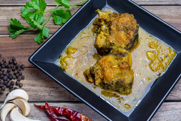 Traditional Bengali cuisine - doi katla (catla fish curry with yogurt) in a black plate with garlic, dry red chilli, black pepper and coriander leaves on wood background.