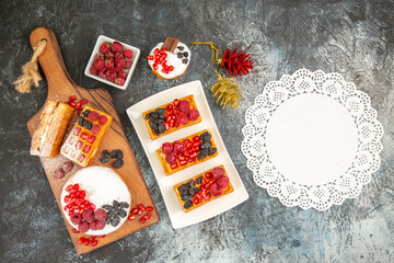 top view different cakes on white rectangular plate on cutting board xmas toys bowl of raspberries white lace doily on grey-white background