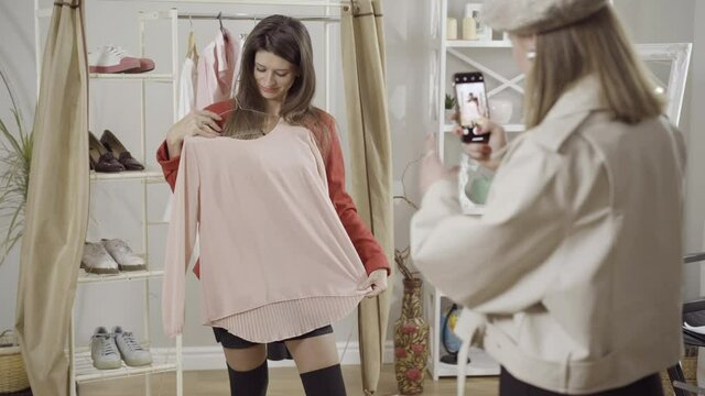 Positive young woman posing with stylish pink pullover in atelier as friend taking photos on smartphone. Happy Caucasian customer choosing garment indoorsion concept.