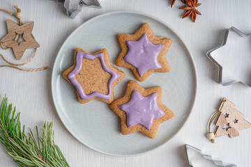 Portion of sweet savory ginger cookies or gingerbread with violet icing served on gray plate cooked for winter holidays on white wooden table with cutters and fir tree. Horizontal, flat lay
