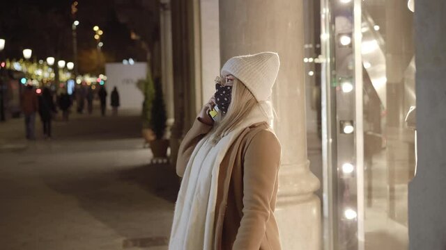 Young blonde woman with a mask in front of a store window talking on the phone and laughing, in a city at night, with lights in the background. She hangs up and leaves the scene. Winter atmosphere.
