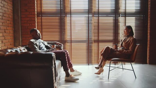 Zoom out of Afro-American businessman sitting on sofa in loft office, drinking to go coffee and speaking with young Asian female colleague