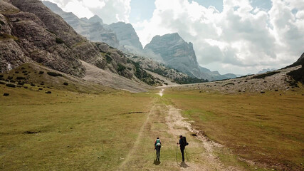 Fototapeta na wymiar A couple hiking along a pathway leading through a valley in Italian Dolomites. High, sharp mountains around. Stony and raw landscape, green meadow. Remote and desolate place. Freedom of exploration