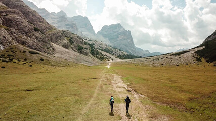 Fototapeta premium A couple hiking along a pathway leading through a valley in Italian Dolomites. High, sharp mountains around. Stony and raw landscape, green meadow. Remote and desolate place. Freedom of exploration